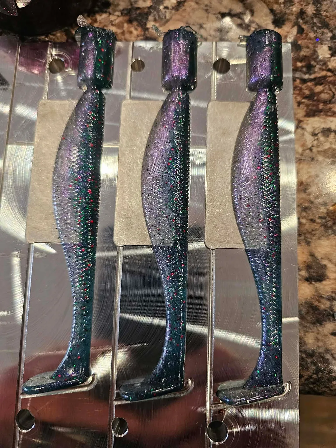 Three fresh purple and blue swimbaits with red and green flake still sitting in an open aluminum mold